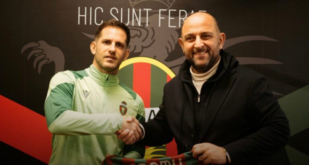 A football player in a green training jersey shakes hands with a man in a black coat, both smiling in front of a graphic backdrop featuring the phrase 'HIC SUNT FERAE' and a colorful logo.