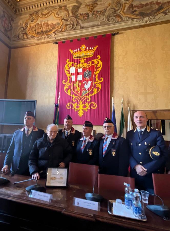 A group of six men in formal attire poses in a decorated room with a large banner in the background. The men, including an elderly man in a black coat, are surrounded by flags and a table with documents and water bottles.