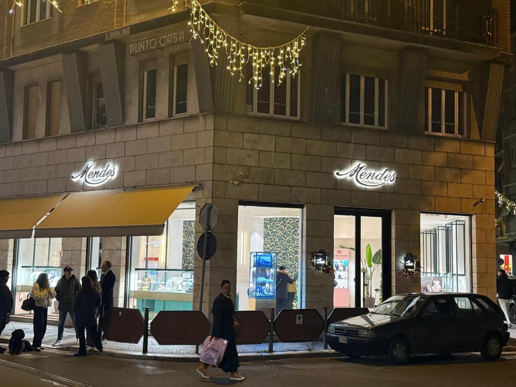 L'ingresso del negozio Mendes illuminato, con persone in fila all'esterno, situato in una strada durante la sera.