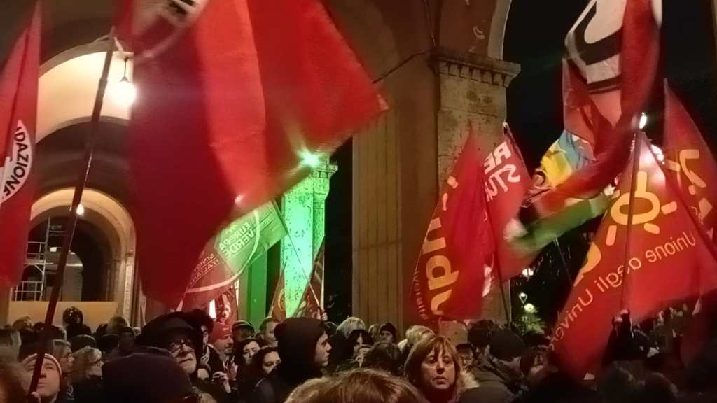 Manifestazione con bandiere rosse sotto un portico di Perugia, con diversi partecipanti visibili.