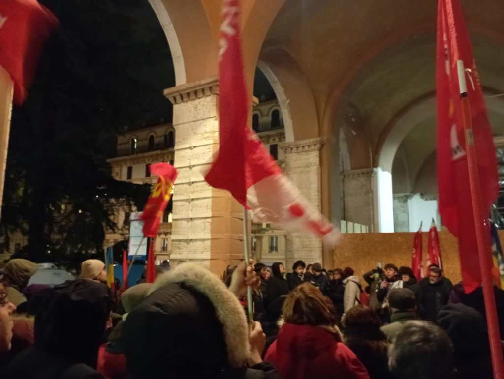 Manifestazione con bandiere rosse sotto un portico in una notte piovosa, con partecipanti che ascoltano un discorso.