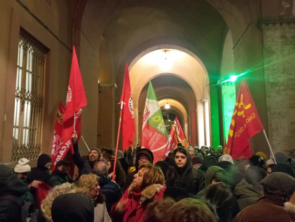 Un gruppo di manifestanti sotto un portico, con bandiere rosse e altri colori, durante una manifestazione a Perugia. I partecipanti indossano abbigliamento pesante per proteggersi dal freddo e dalla pioggia.