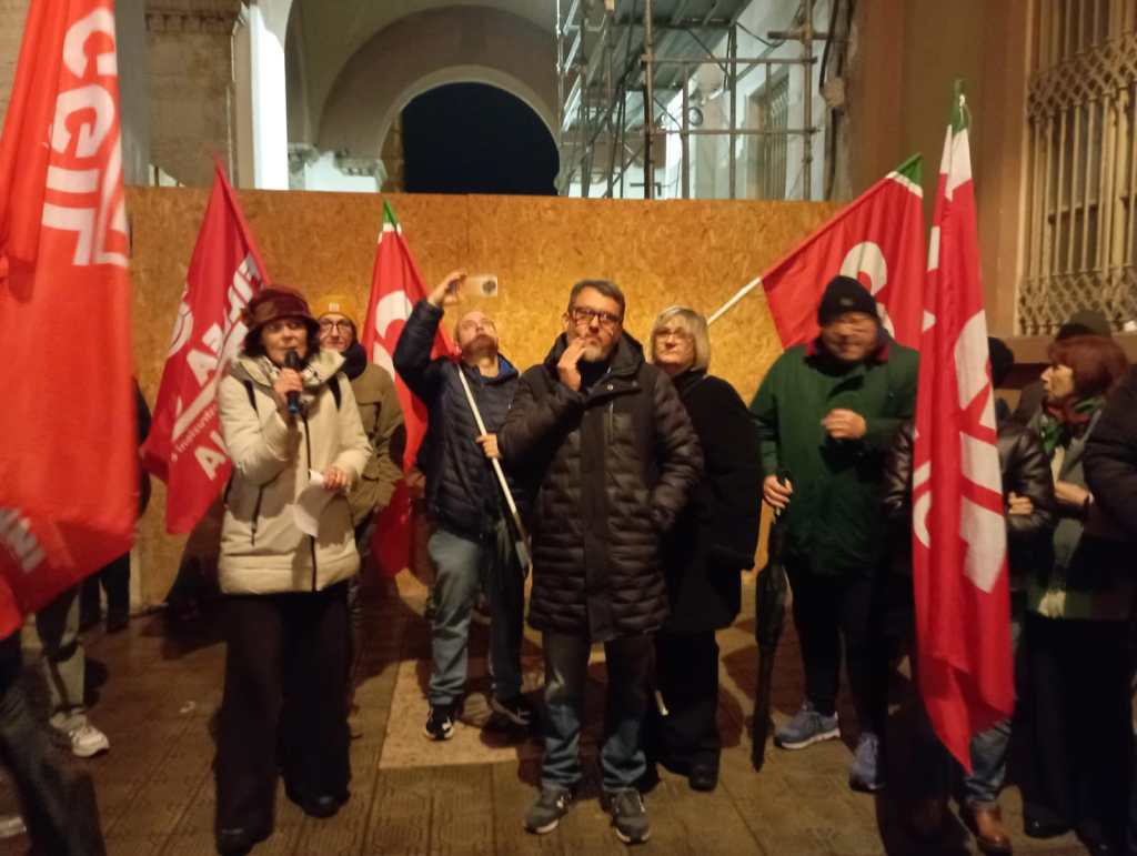 Manifestazione della Cgil a Perugia con partecipanti che reggono bandiere rosse e ascoltano un intervento al microfono.