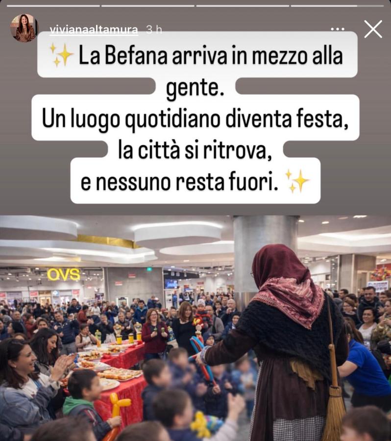 Una figura vestita da Befana interagisce con un gruppo di bambini in un evento festivo in un centro commerciale affollato, con un tavolo decorato sullo sfondo e persone che applaudono.