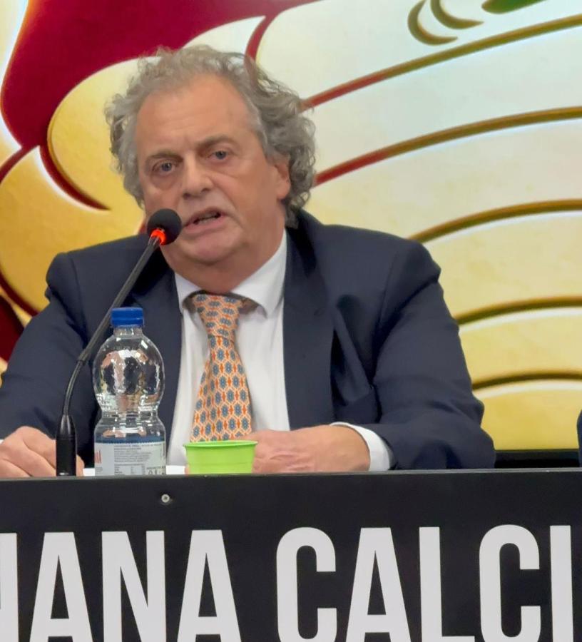 A man with curly hair sits at a podium with a microphone, engaged in a discussion. He is wearing a suit and a patterned tie, with a water bottle and a small green cup on the table in front of him.