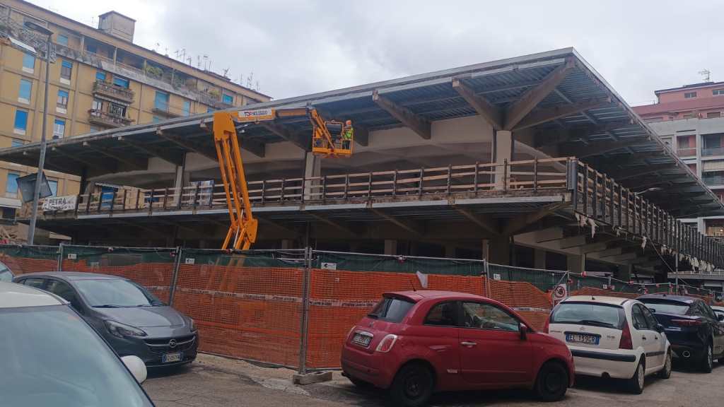 Construction site of a building with scaffolding and workers using a lift, surrounded by parked cars.