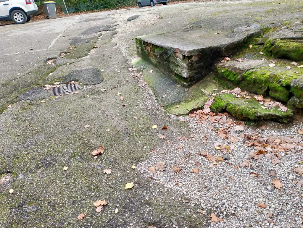 Un'area di parcheggio con pavimentazione irregolare e fatiscente, evidenziando buche e vegetazione incolta, con foglie secche sparse e un gradino danneggiato sul lato.