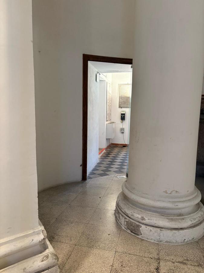 A view through an archway showing a corridor with tiled flooring and a white wall. On the right, a large column is partially visible.