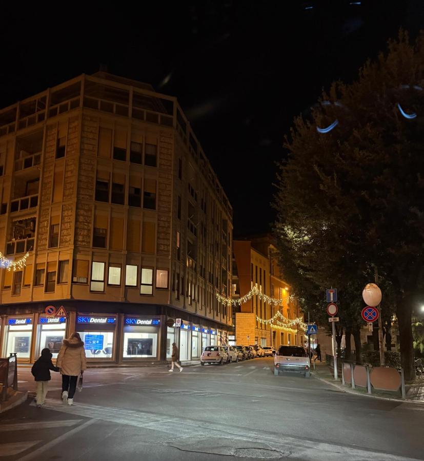 A nighttime street scene featuring a corner building with bright windows and holiday decorations. Two people walk along the sidewalk, and parked cars line the street.