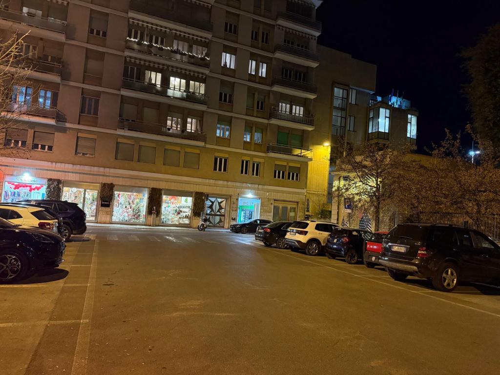 A nighttime view of a city street featuring parked cars on either side, with illuminated buildings and shop windows in the background.