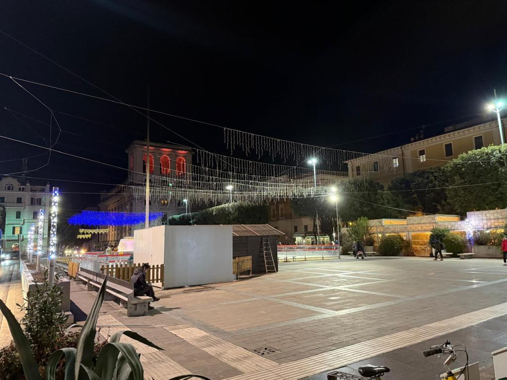 A lit-up square at night, featuring decorative lights hanging above. People are seen walking and sitting on benches, with buildings illuminated in the background.