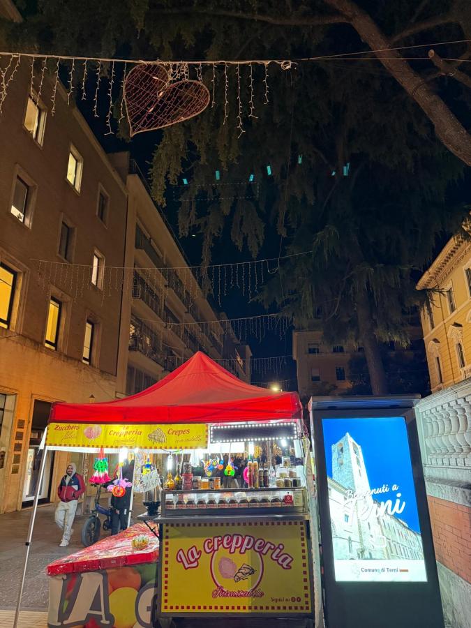 Nighttime view of a vibrant food stall with a red canopy titled 'La Crepperia', offering various colorful treats, set against a backdrop of illuminated buildings and festive decorations.