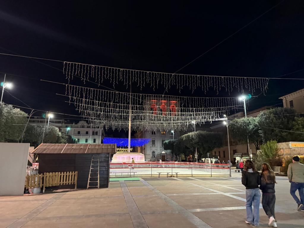 Illuminated street with decorative lights overhead, a fountain in the background, and people walking in a plaza at night.