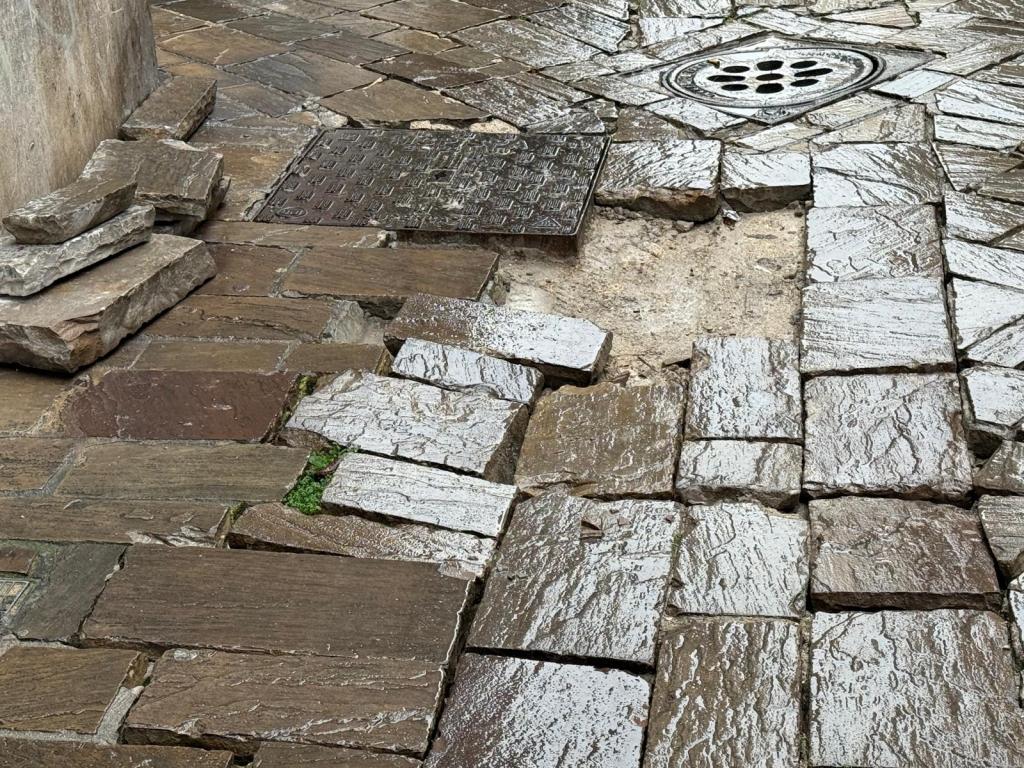 Wet stone pavement with uneven tiles and a drain cover, showing patches of dirt and grass.