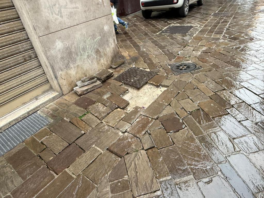 Wet cobblestone street with irregular paving stones, showing a drain cover and a small stack of stones beside a building.
