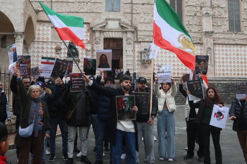 A group of protesters holding signs and flags, advocating for Iran, in a public square.