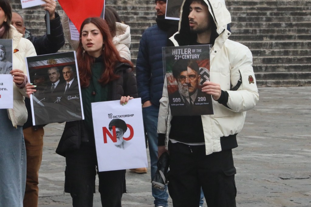 A group of protesters holding signs at a rally. One person is displaying a poster with the text 'Hitler of the 21st Century' alongside an image, while another holds a sign that says 'NO TO DICTATORSHIP'.