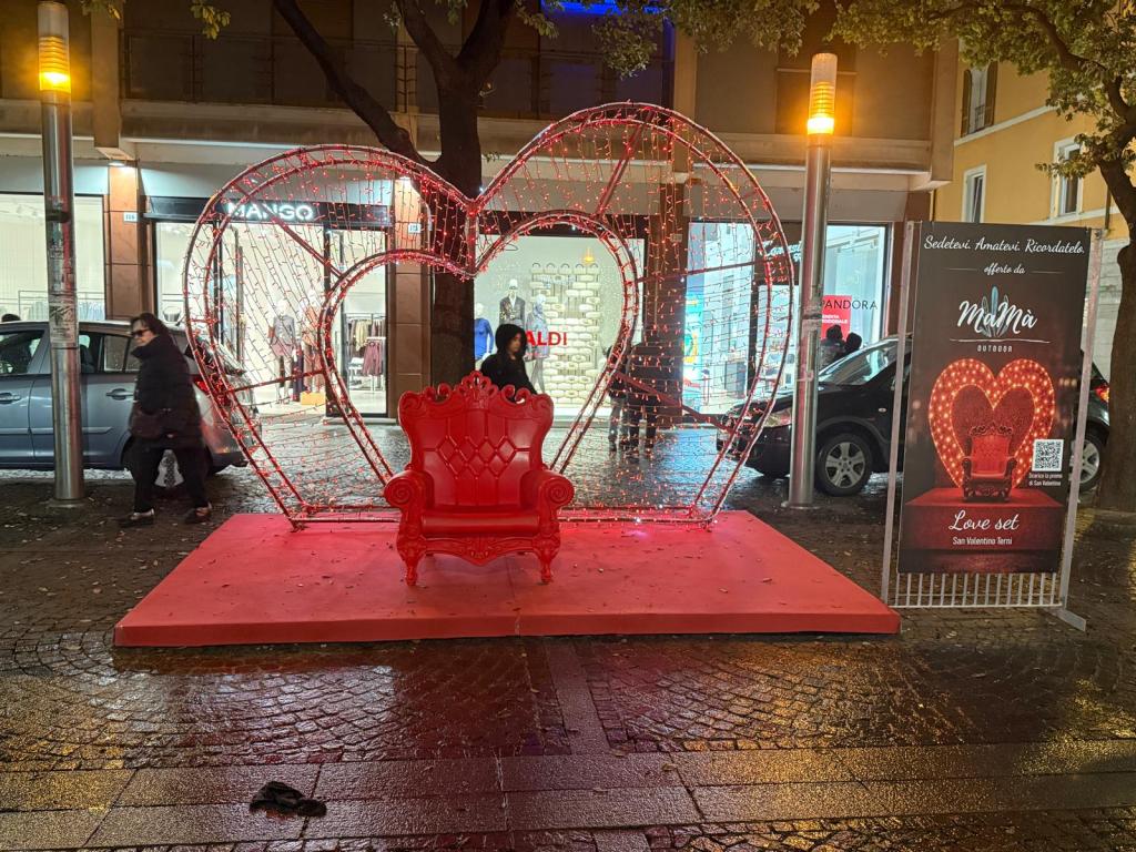 A red velvet throne with an illuminated heart backdrop, set in a bustling street, promoting a Valentine's Day event.