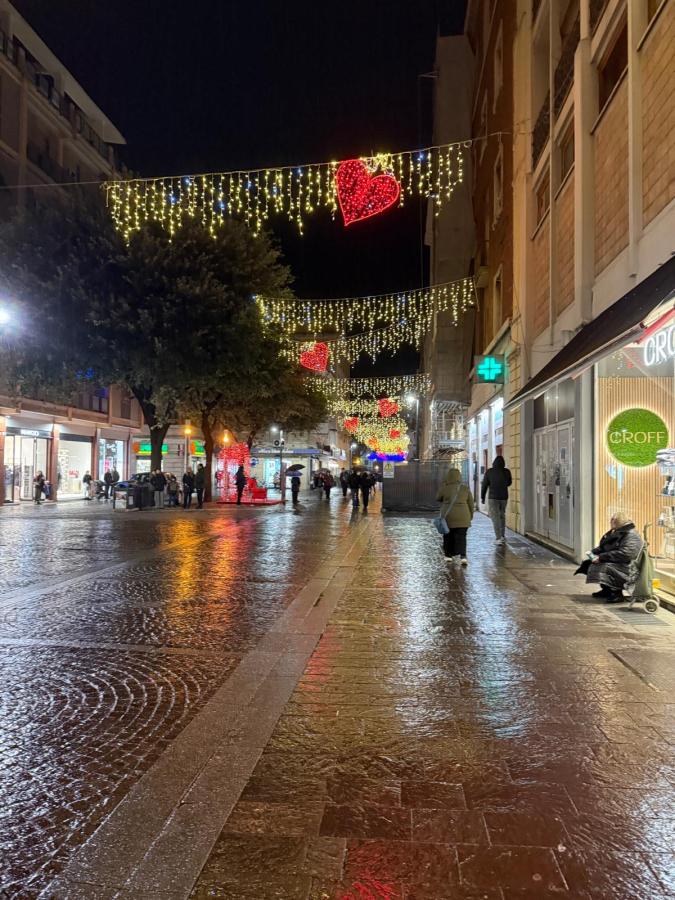 A brightly lit street at night decorated with heart-shaped lights, reflecting on a wet pavement, with people walking and shops along the sides.