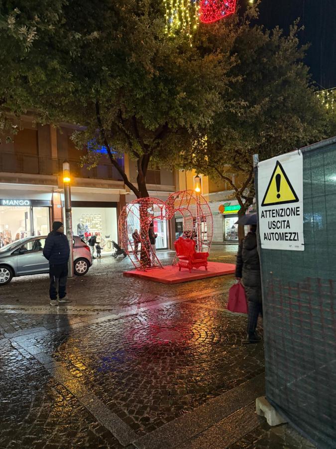 A vibrant public square at night featuring a decorative heart-shaped structure with a red chair inside, surrounded by shoppers and festive lights. Rain reflects on the cobblestone pavement, and a caution sign for vehicles is visible nearby.