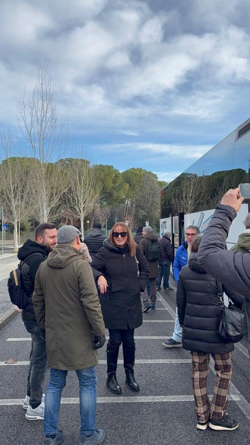 A group of people standing in a line next to a bus, with some engaged in conversation. The scene is set outdoors on a cloudy day with trees in the background.