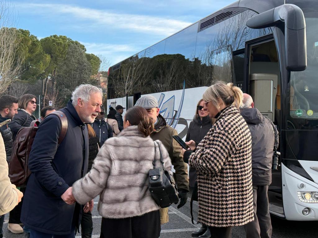 A group of people interacting near a bus parked in an outdoor setting, with some individuals engaged in conversation and others using mobile devices.