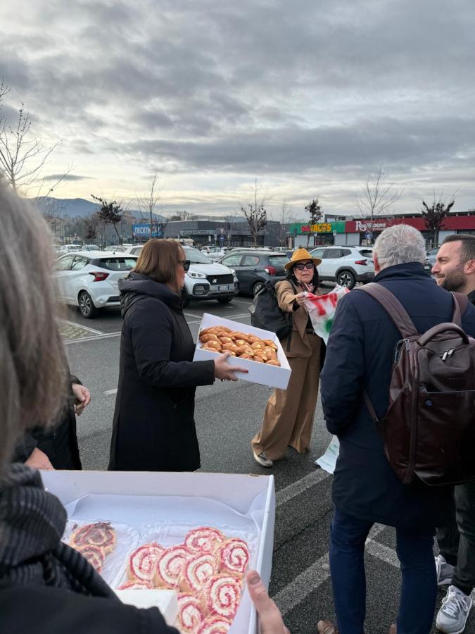 A group of people in a parking lot, with one woman holding a box of pastries while another woman gestures with a flag. Several cars are parked in the background under a cloudy sky.