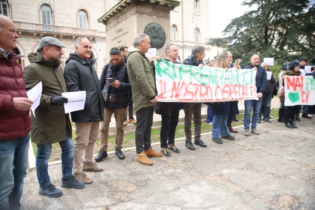 A group of people gathered outdoors holding banners and signs during a protest. They are dressed in winter clothing, standing in front of a building with trees in the background.