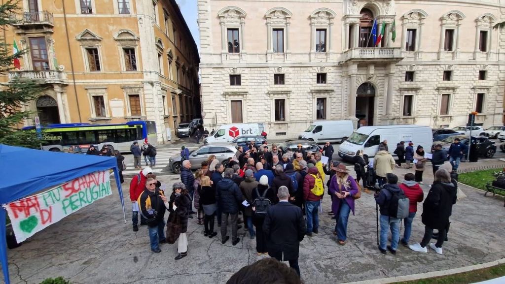 A crowd gathers in front of a historic building, with signs and banners visible, during a demonstration in an urban setting.