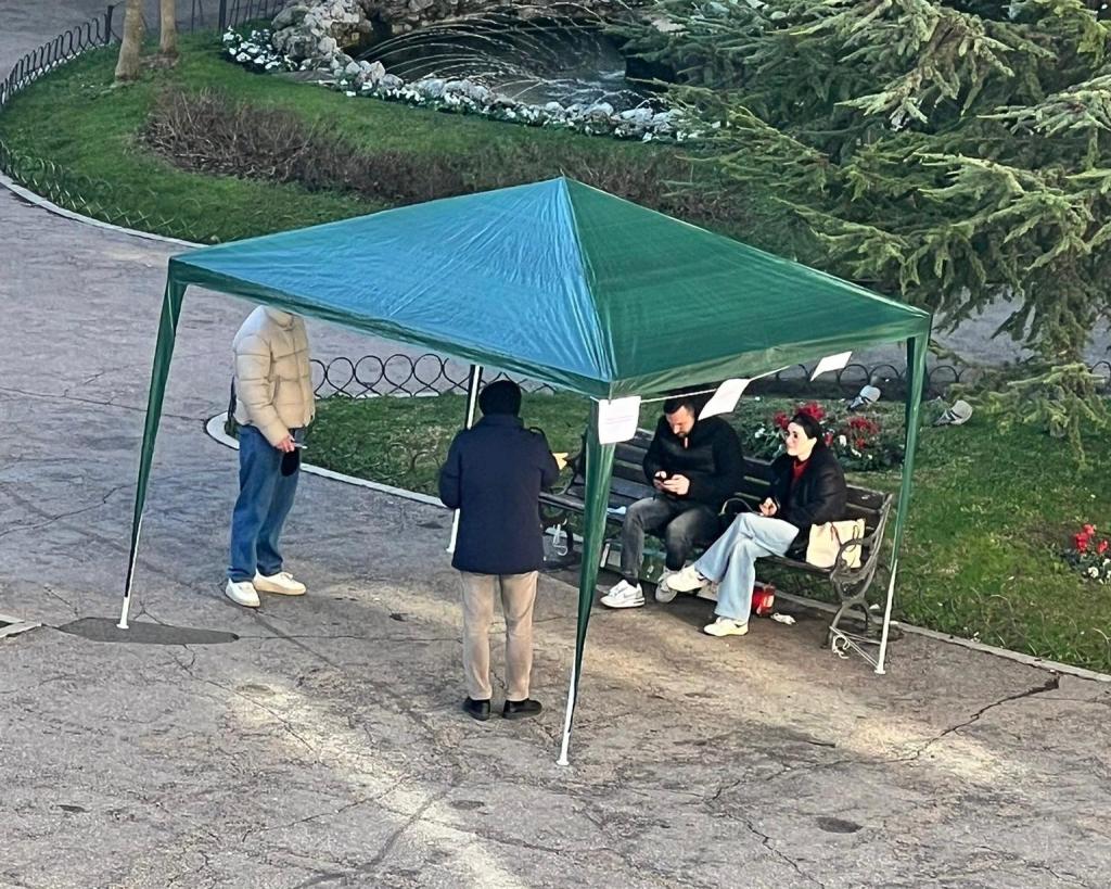 People gathered under a green tent in a park, with one person standing and interacting while others sit on a bench checking their phones.