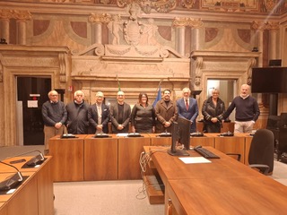 Group of eight individuals standing in a formal setting with a large decorative backdrop and wooden furniture, likely in a government or conference room.
