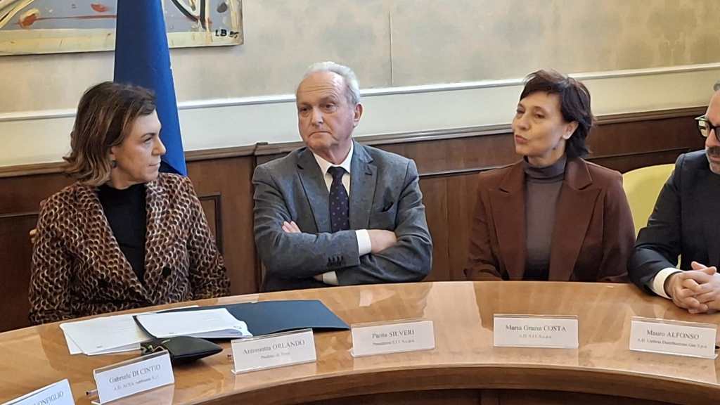 A group of four professionals seated around a table during a meeting. They appear to be engaged in discussion, with two women and two men present. A blue flag is visible in the background.