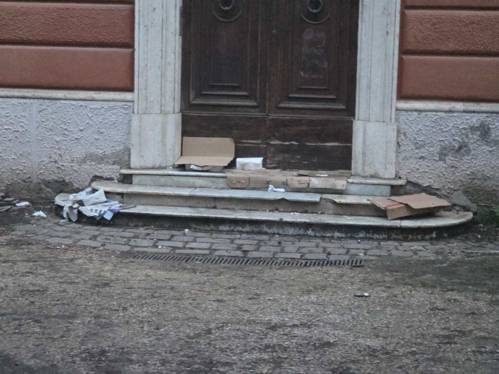 A stoop with steps leading to a wooden door, cluttered with cardboard boxes and litter.