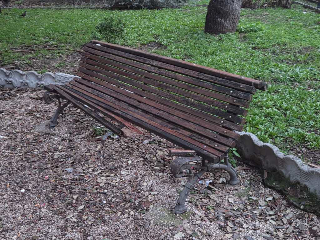 Abandoned wooden park bench placed on gravel ground surrounded by grass and leaves.