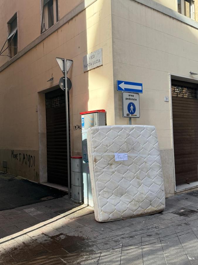 A discarded mattress propped against a wall on a city street corner, with street signs indicating 'Vico Santa Lucia' and a pedestrian area.