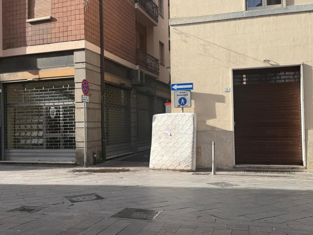 An urban street scene featuring a discarded mattress leaning against a wall, with a pedestrian area sign and a closed storefront in the background.