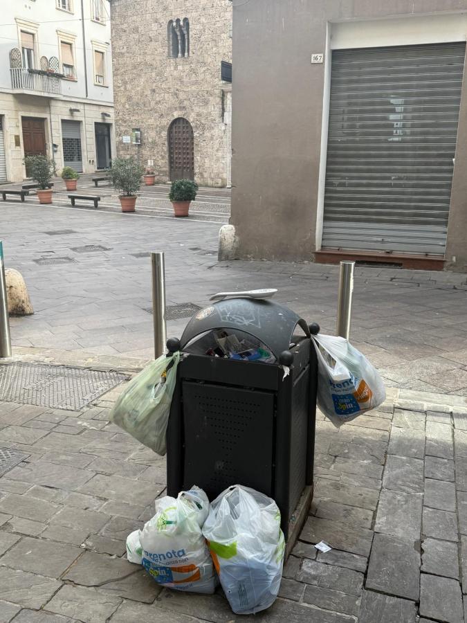 A black trash bin overflowing with garbage bags, situated on a cobblestone street next to a building with a stone facade and closed shutters.