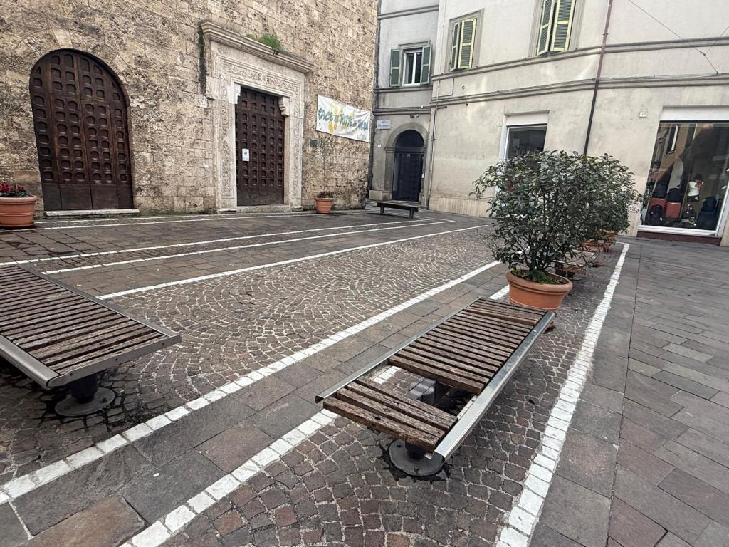 A cobblestone plaza featuring several wooden benches, potted plants, and large wooden doors in the background, with a banner hanging on the wall.