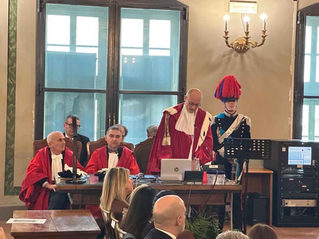 A courtroom scene with several judges in red robes seated at a table, one judge standing and speaking. A guard in ceremonial uniform stands to the side. The room has large windows and an ornate chandelier.