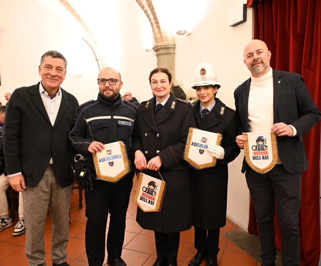 Un gruppo di quattro persone posano insieme, tre di loro in uniforme e una in abiti casual, mentre mostrano dei manifesti legati all'evento della Befana dell'ASI.