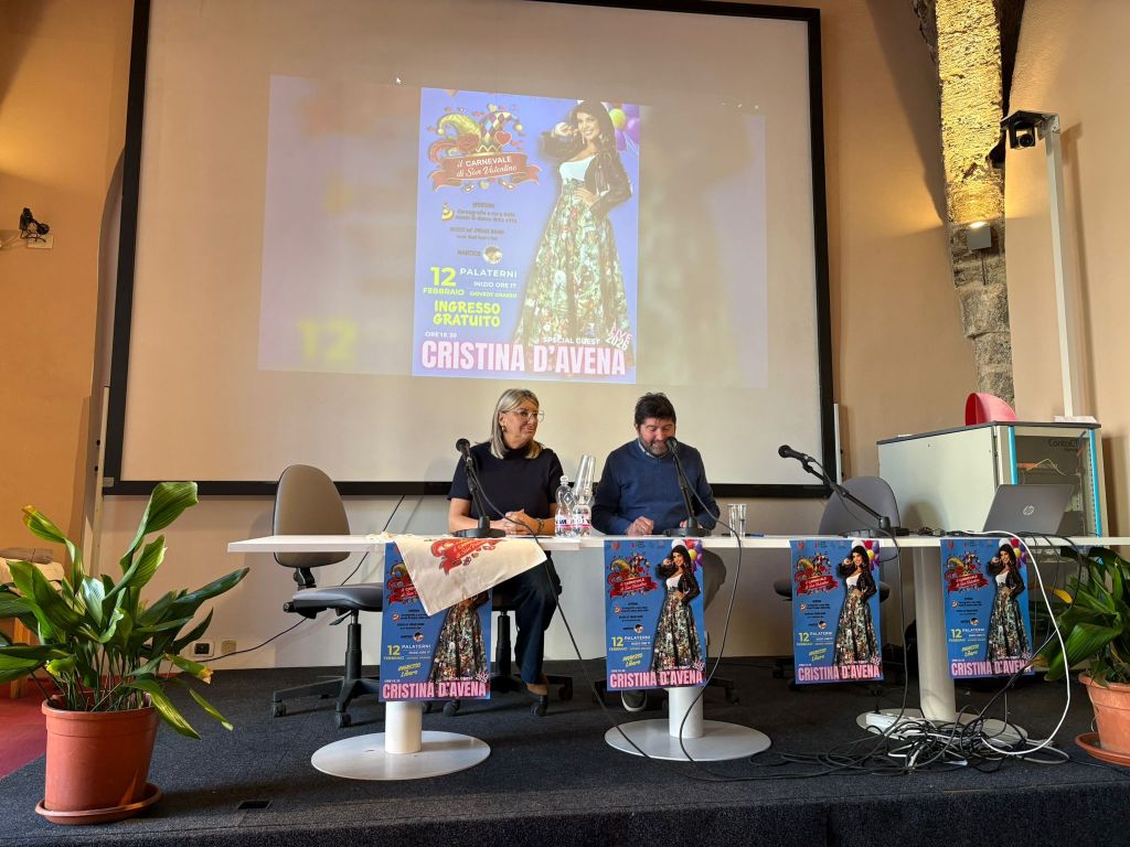 A press conference featuring two presenters seated at a table with promotional posters for Cristina D'Avena and an event announcement on a large screen behind them.