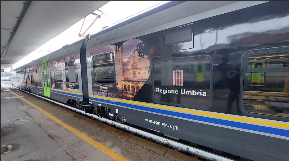 Treno Minuetto della Regione Umbria in sosta alla stazione, con livrea dedicata alla promozione turistica e immagini iconiche del territorio.