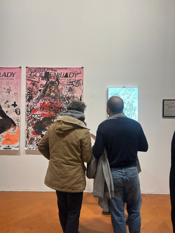 Two people observing artwork in an art gallery, with colorful posters displayed on the wall and a lighted piece of art beside them.