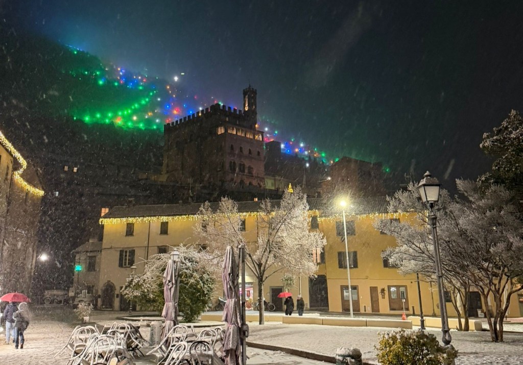 Piazza di Montone decorata con neve e luci colorate durante la notte, con persone che passeggiano sotto la neve.