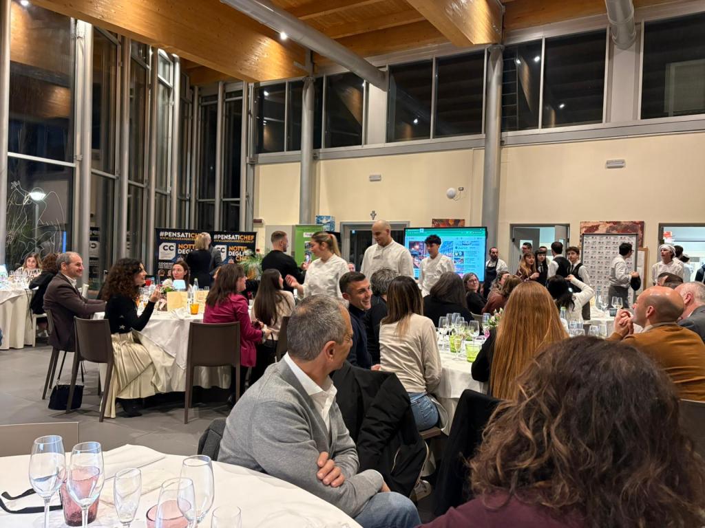 A busy restaurant interior with guests seated at tables, enjoying a meal. Several staff members in white uniforms are serving food. The atmosphere appears festive with large windows, soft lighting, and elegantly set tables.