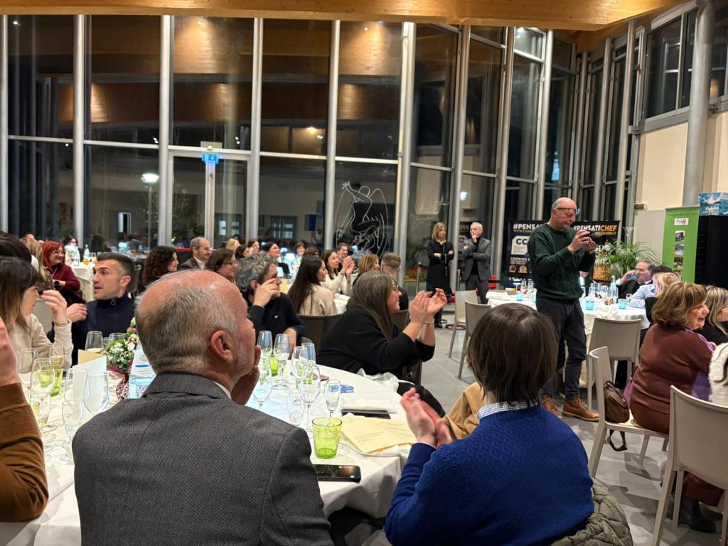 A lively gathering in a spacious venue with large windows, featuring several tables filled with guests engaged in conversation and applauding. A speaker stands and addresses the audience, while others react positively and enjoy the event.