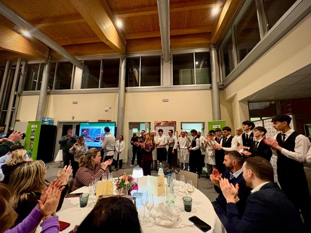 A group of people applauding at a gathering, with servers in formal attire and guests seated at tables in a well-lit venue.