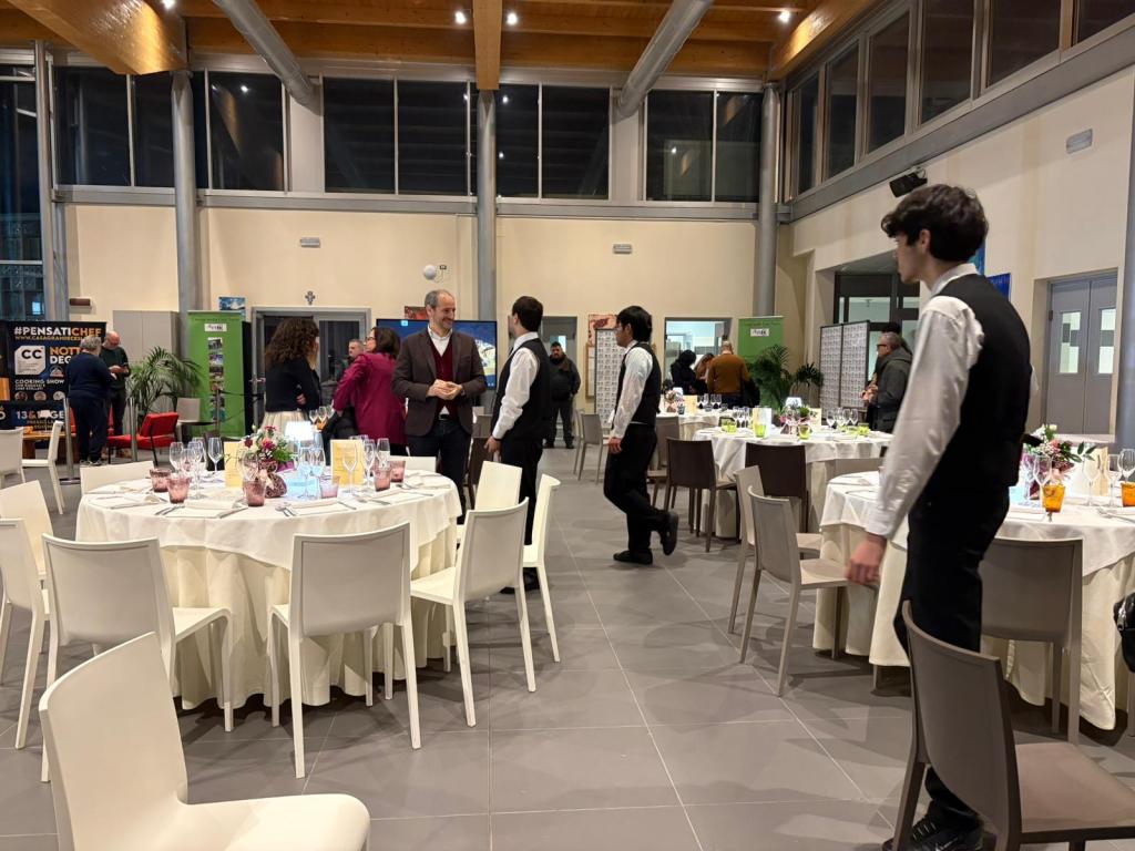 A spacious dining area set for an event, featuring round tables with white tablecloths and decorative centerpieces. Attendees are mingling, and staff in uniforms are serving drinks. The venue has large windows and a modern design.