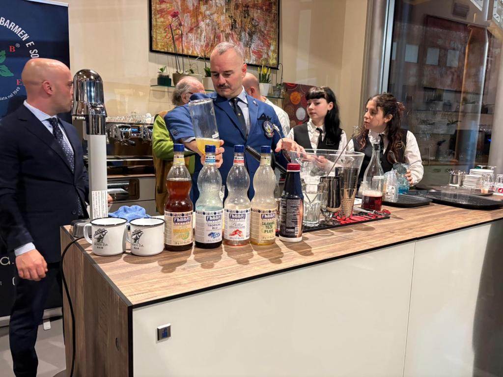 A group of bartenders working behind a bar, preparing drinks. Various bottles of mixers are lined up on the counter, and the bartenders are engaged in a mixology demonstration.