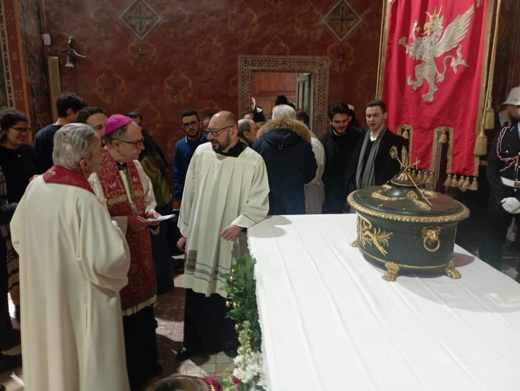A group of people including clergy members in ceremonial attire engages in conversation near a decorative casket on a table, set within an ornate interior space.